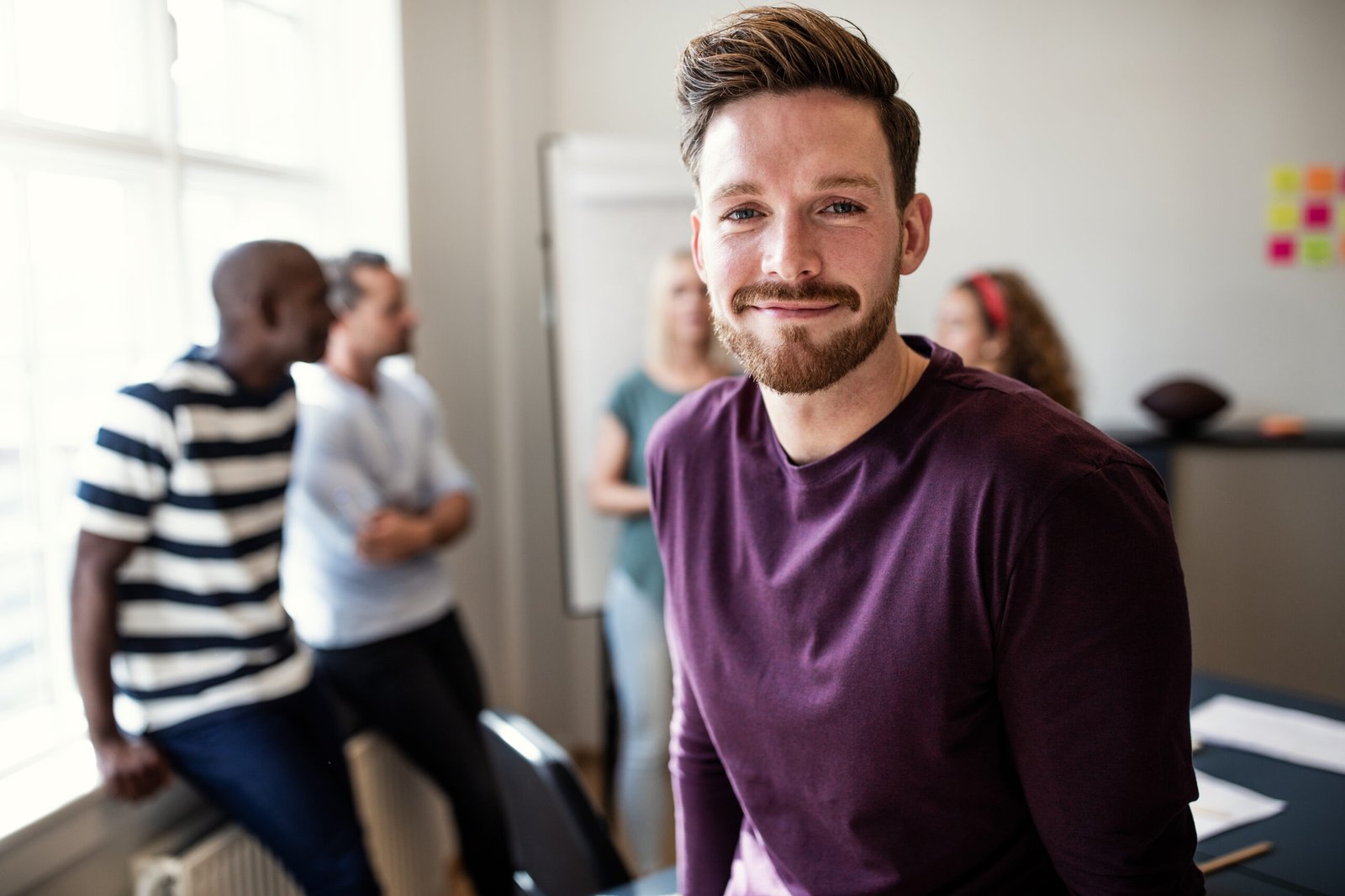Young designer smiling confidently in an office after a presentation Smiling young designer standing in an office after a boardroom meeting with colleagues talking together in the background